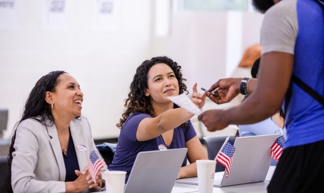 Two women are working at a polling place helping a man receive his ballot