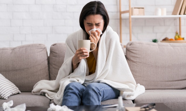 Young woman with a cold sitting on a couch covered in a blanket, blowing her nose and drinking hot tea
