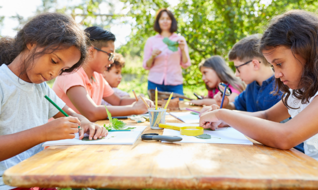 Group of elementary age children working on school projects outside at a picnic table on a sunny day.