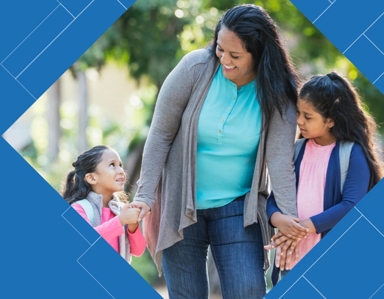 A middle-aged Hispanic woman walks her two happy-looking young daughters to school while they hold her hands