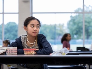 Elementary-age girl sits alone in a school cafeteria looking sadly at others off camera.