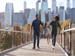 Two parents lift a small child up by the hands between them as they walk across a bridge in a large city.