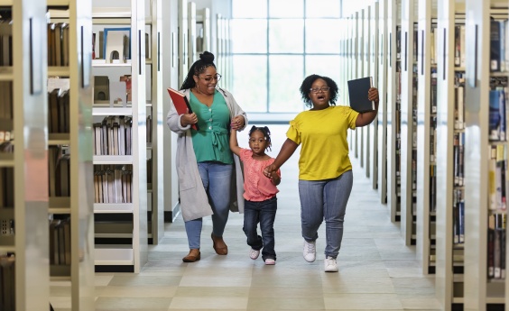 Two women and a young girl walk hand in hand in a library, excited about finding good books