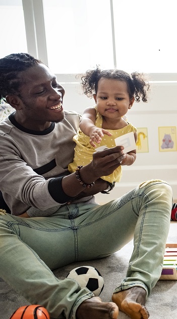 A young black father plays inside with his toddler daughter while both look very happy.
