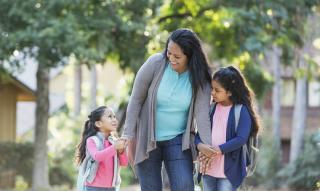 A middle-aged Hispanic woman walks her two happy-looking young daughters to school