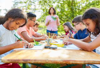 Group of elementary age children working on school projects outside at a picnic table on a sunny day.