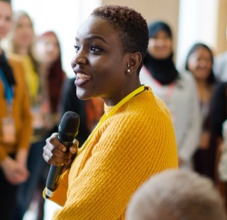A young women in a bright yellow sweater speaks on a microphone in a large group of people
