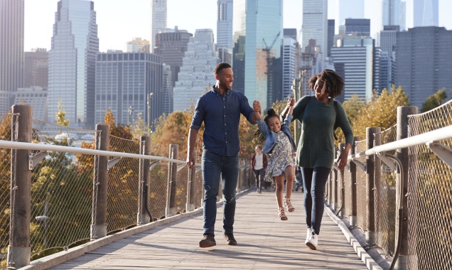 Two parents lift a small child up by the hands between them as they walk across a bridge in a large city.