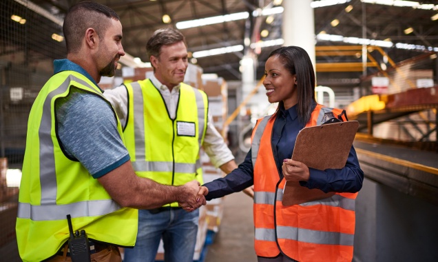 Two people wearing neon work vests and shaking hands in a modern factory.